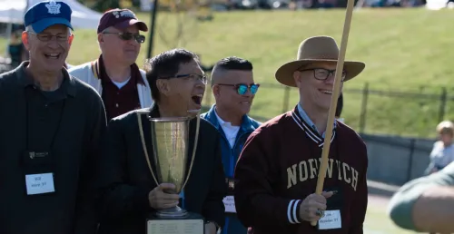 five men, most smiling, led by one in front wearing hat with sweatshirt reading Norwich, and second carrying a trophy