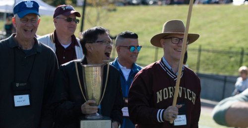 five men, most smiling, led by one in front wearing hat with sweatshirt reading Norwich, and second carrying a trophy