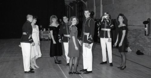 four couples, men dressed in uniform, women in formal event dresses, on parquet dancefloor
