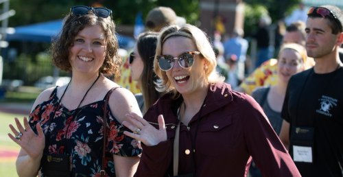 two women alumnae of Norwich University, both wearing sunglasses (at left on her forehead), waving.