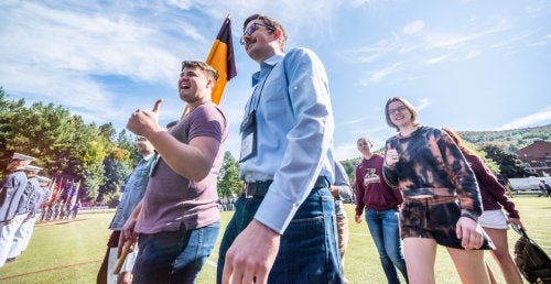five individuals in casual clothing marching on a football field. Man leading the group holding a flag / guidon.