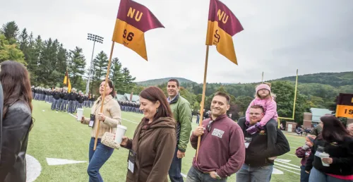 men, women and father with child on his shoulders, walk on football field, with a woman (left) and a man (right) holding flags / guidons.