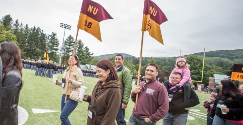 men, women and father with child on his shoulders, walk on football field, with a woman (left) and a man (right) holding flags / guidons.