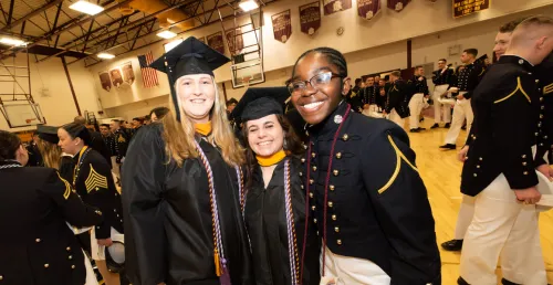 Three people wearing graduation caps and gowns standing next to a person in a dark uniform with gold trim in a gym.