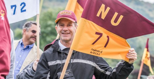 man in red cap and black & gray jacket holds a guidon flag with "NU 71 Compliments of the N.U.A.A." written on it. Photo of Bob Gillespie '71, at the Norwich Univ. Homecoming, 2014.