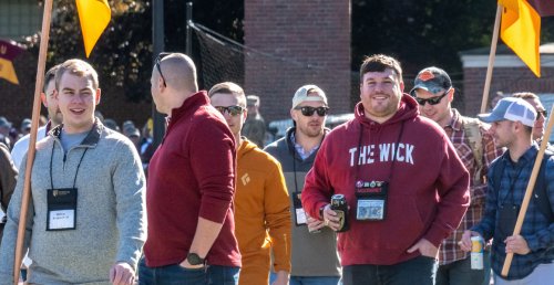 Seven individuals in casual attire walk forward, several holding up maroon and gold flags (guidons).
