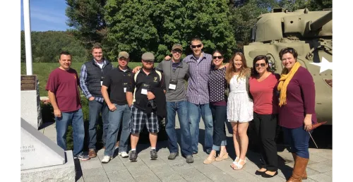 A group of individuals in casual dress stand together with a part of a tank visible behind them at right.