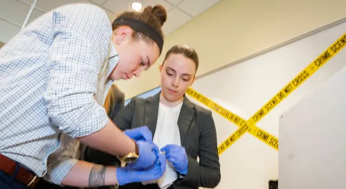 Two students wearing gloves work together on a hands-on forensic-style lab activity marked with caution tape.
