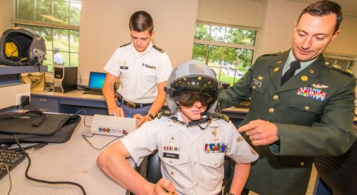 Cadet wears a flight helmet as an officer adjusts equipment in a classroom.
