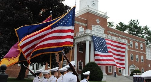 Cadets carry American and Norwich flags in formation in front of Jackman Hall, where a large American flag hangs from the building’s columns.