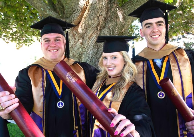 Norwich online graduate students in their graduation gowns with their diplomas.