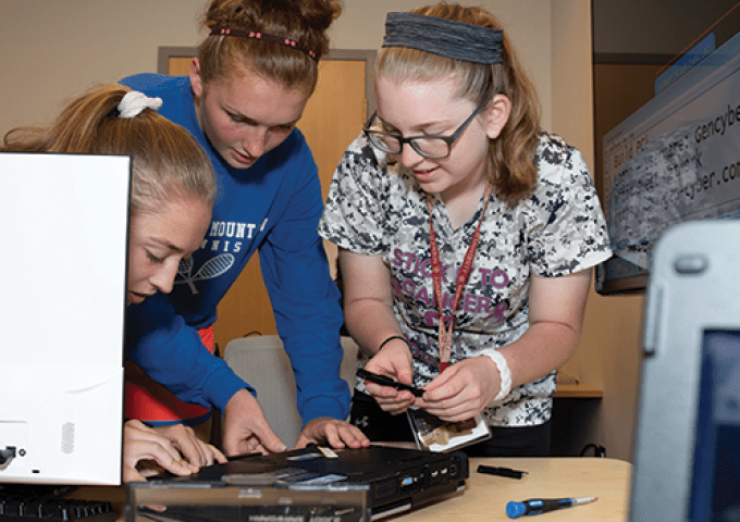 Three high schoolers work together on hardware surrounded by computers.
