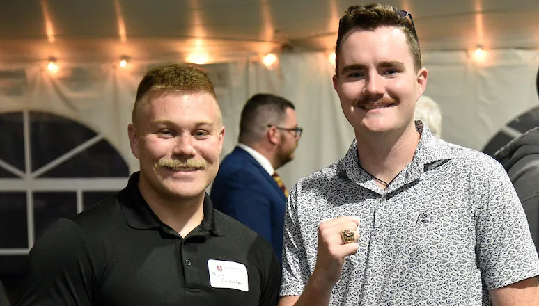 two men standing in a tented event, the fellow on the right holding up his hand bearing a ring.