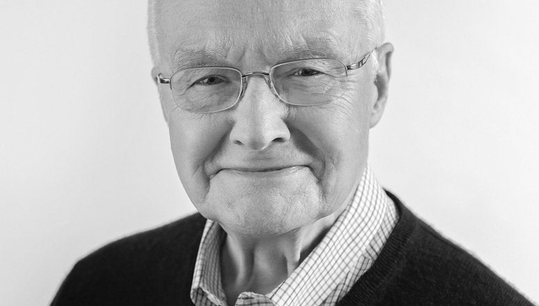 Person with short gray hair and glasses wears a dark sweater over a checkered shirt against a plain background.