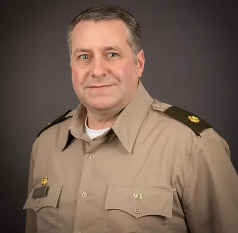 A person in a tan military-style uniform stands against a dark background, facing slightly to the left.