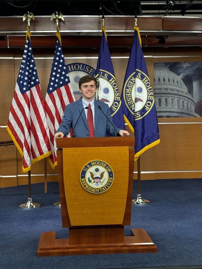 Spencer Rada stands behind a U.S. House of Representatives podium with a set of flags in the background.