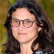 A woman with shoulder-length dark brown hair, wearing tortoiseshell glasses and a patterned blouse, standing outdoors with a softly blurred green and brown background.