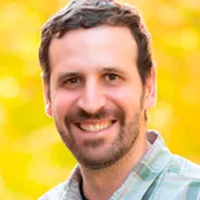 A man with short, wavy brown hair and a beard, wearing a plaid shirt, standing outdoors with a blurred golden-yellow background.