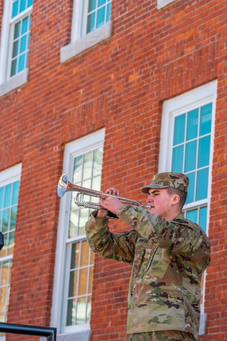 A mid-range shot of a cadet standing on the stairs of Jackman Hall with a bugle raised.