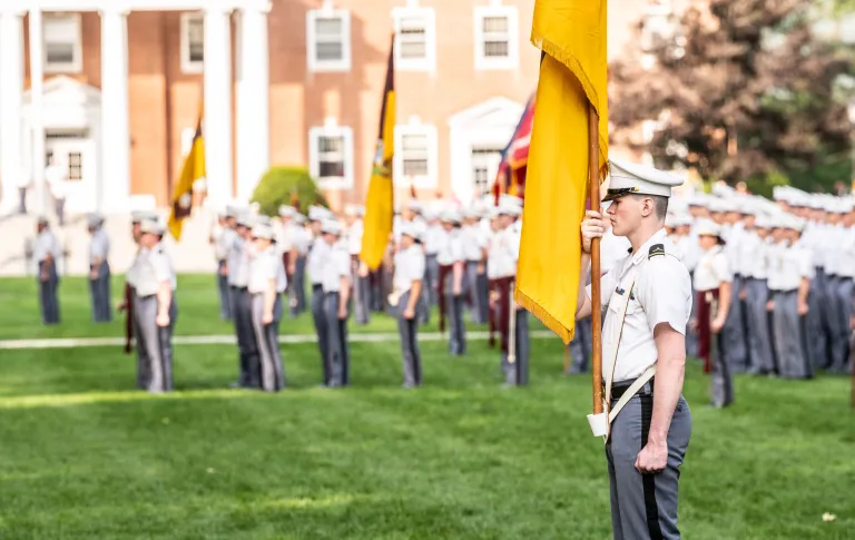 A cadet stands on the Upper Parade Ground during a Review with Retreat, and is holding a flag in his right hand. There are other groups of cadets in the background.