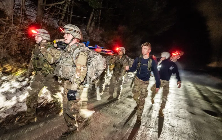 A group of cadets walk along a road with snow on the shoulder in the dark with lights shining from behind. The majority of the group are working together on the left while two leaders are on the right.