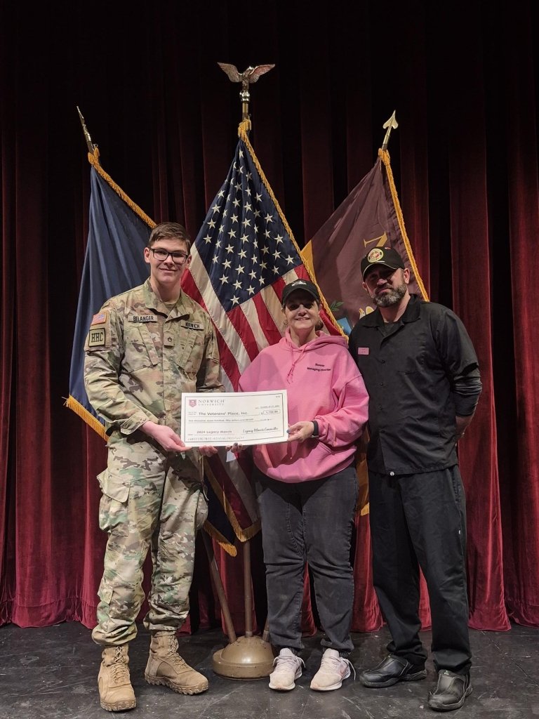 Three people are standing on stage between the US flag and other military banners, one in military uniform and two in civilian clothing, holding a certificate together.