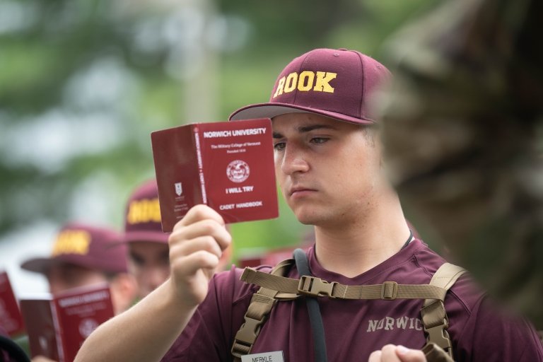 A male Rook is in maroon shirt and hat, standing at attention with his Rook Book directly held in front of his face.