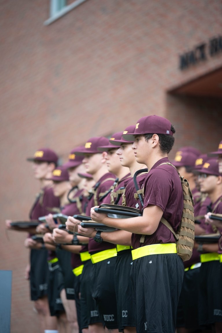 A group of Rooks stand together in maroon shirts and hats with black shorts, holding shirts in front of them.