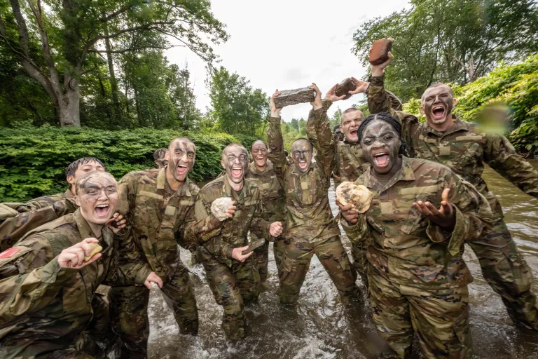 A group of cadets smile and yell towards the camera. They are standing in the Dog River having completed the Dog River Run and are holding their rocks.