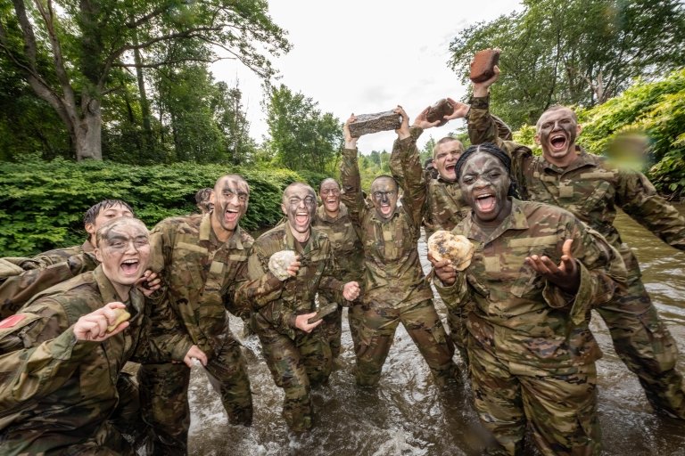 A group of cadets smile and yell towards the camera. They are standing in the Dog River having completed the Dog River Run and are holding their rocks.