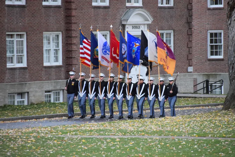 Cadets march in a line along the Upper Parade Ground carrying flags of each branch.