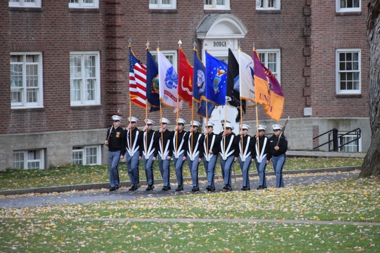 Cadets march in a line along the Upper Parade Ground carrying flags of each branch.