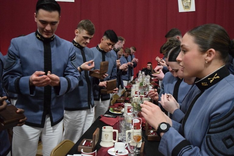 Cadets in uniform at a formal dining event, examining rings at a table decorated with red linens.