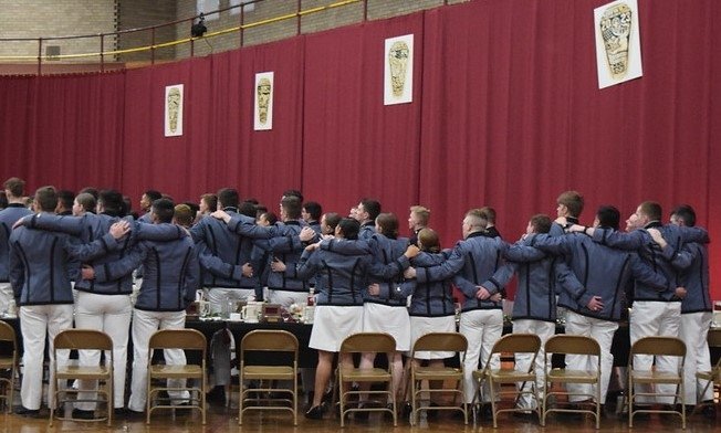 A group of individuals in matching uniforms of blue tops and white bottoms stands with arms around each other's shoulders in a gymnasium, facing a stage with red curtains and banners.