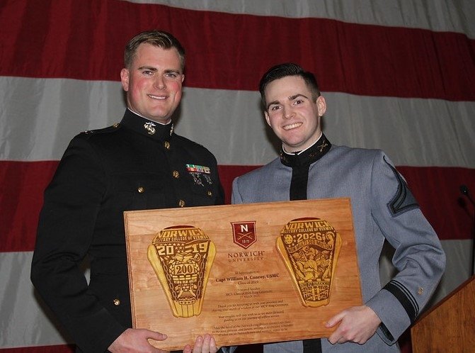 Two individuals in formal attire holding an award plaque at a Norwich University event. The plaque commemorates achievements for the years 2019 and 2026.