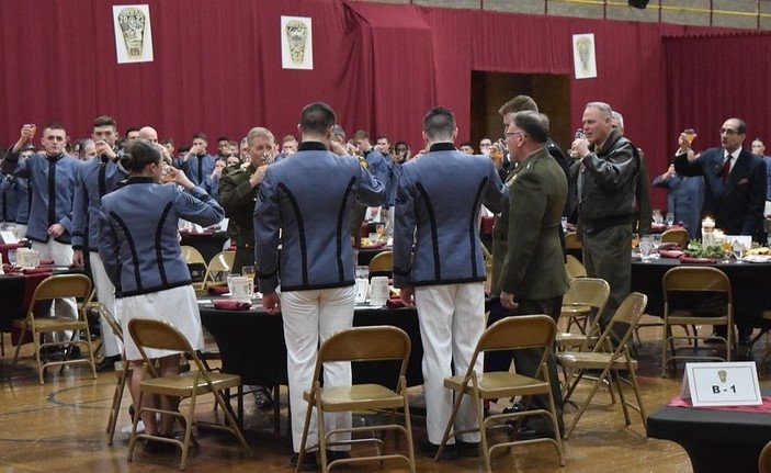 Cadets in formal uniforms participating in a toast at a Norwich University military banquet in a hall decorated with banners and shields.
