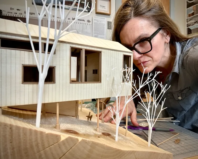 Person examining an architectural model of a modern house with white tree models on a wooden table.