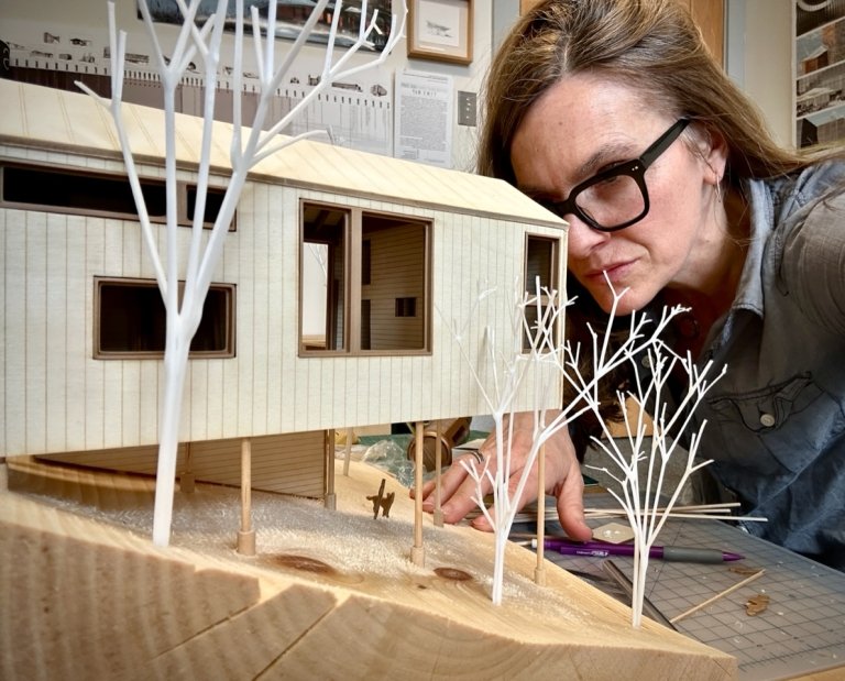 Person examining an architectural model of a modern house with white tree models on a wooden table.