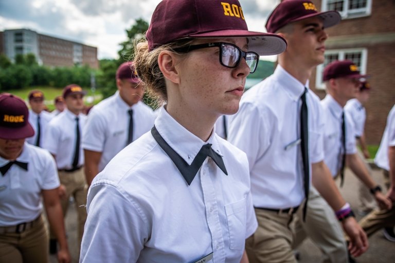 A close-up of a young female rook in glasses, Rook hat, and uniform, marching alongside other rooks on campus.