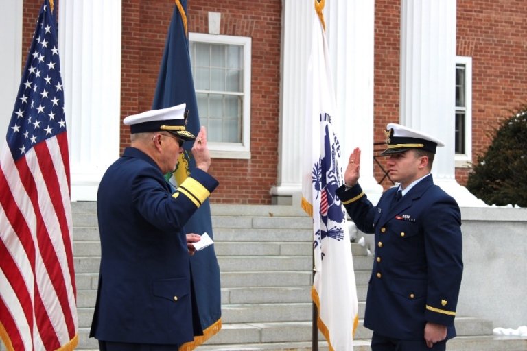 Military ceremony with two naval officers in dress uniform saluting each other in front of a brick building with American flags.