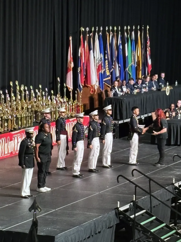 A group of individuals in military uniforms are standing onstage at a formal awards ceremony with a variety of international flags and numerous trophies displayed behind them. The setting includes a stage with a dark curtain backdrop and an audience in the foreground.