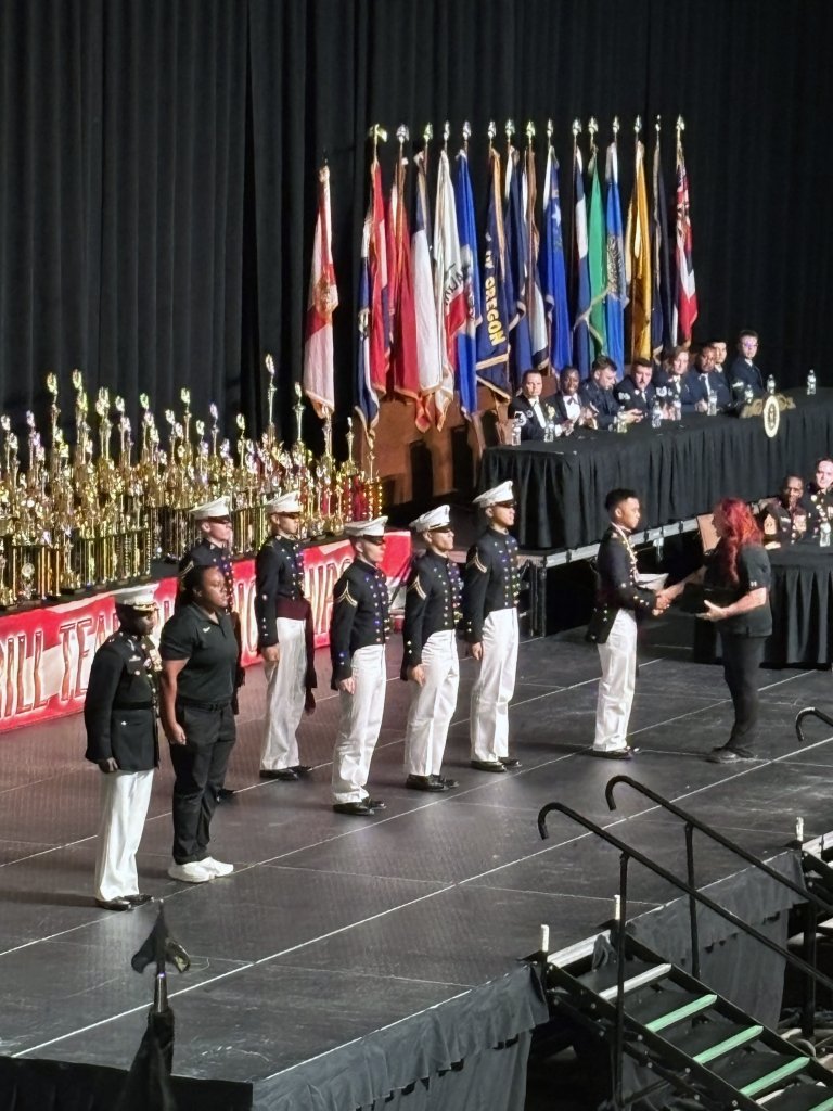 A group of individuals in military uniforms are standing onstage at a formal awards ceremony with a variety of international flags and numerous trophies displayed behind them. The setting includes a stage with a dark curtain backdrop and an audience in the foreground.