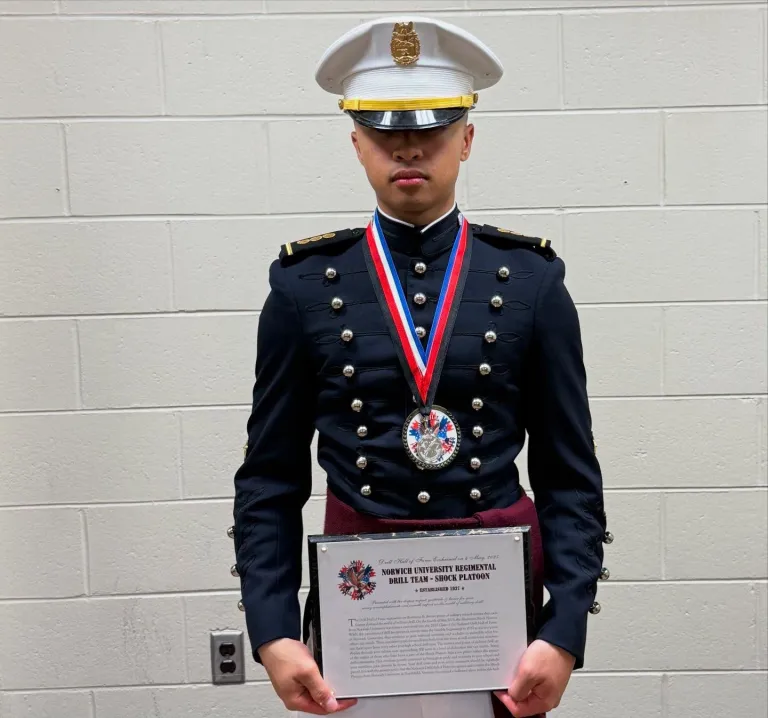 Person in Norwich University Corps of Cadets dress uniform holding a medal and certificate.