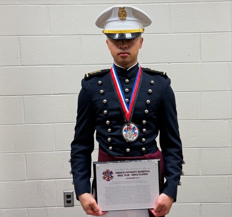 Person in Norwich University Corps of Cadets dress uniform holding a medal and certificate.