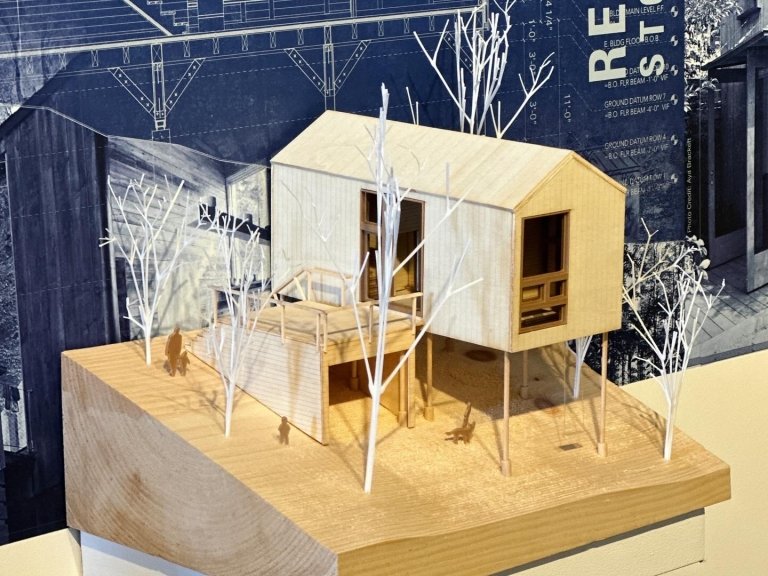 A detailed architectural model of a small, modern house surrounded by trees, displayed against a backdrop featuring blueprints and construction plans.