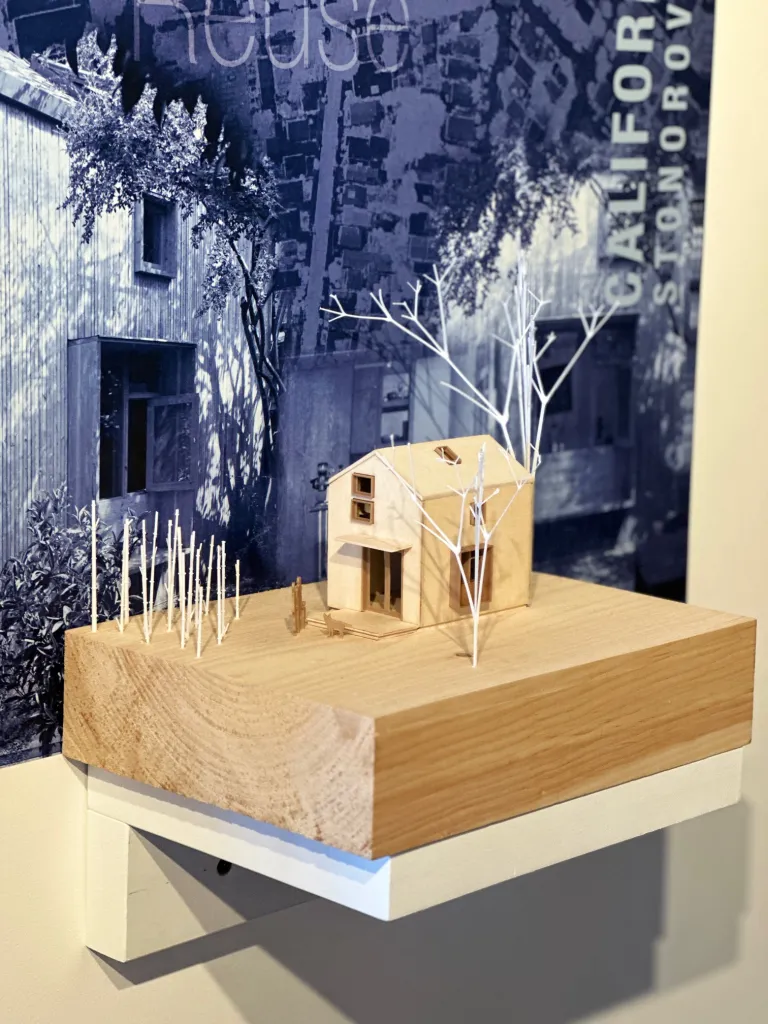Model of a small wooden house and trees displayed on a wood block with a large grayscale photo background featuring architectural imagery.