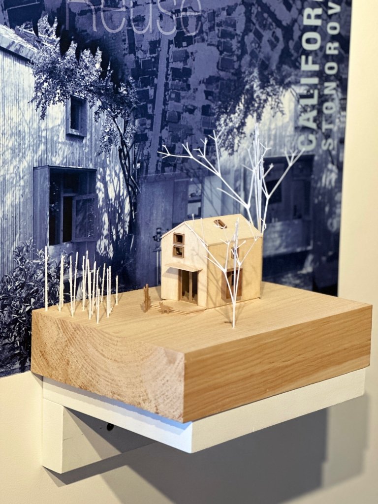 Model of a small wooden house and trees displayed on a wood block with a large grayscale photo background featuring architectural imagery.