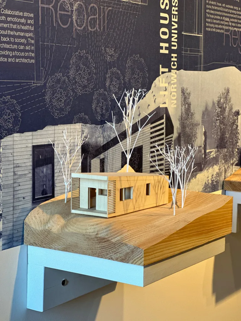 Architectural model featuring a detailed scale model of a minimalist house with surrounding trees displayed on a wooden platform, against a background of informational panels with text and diagrams.