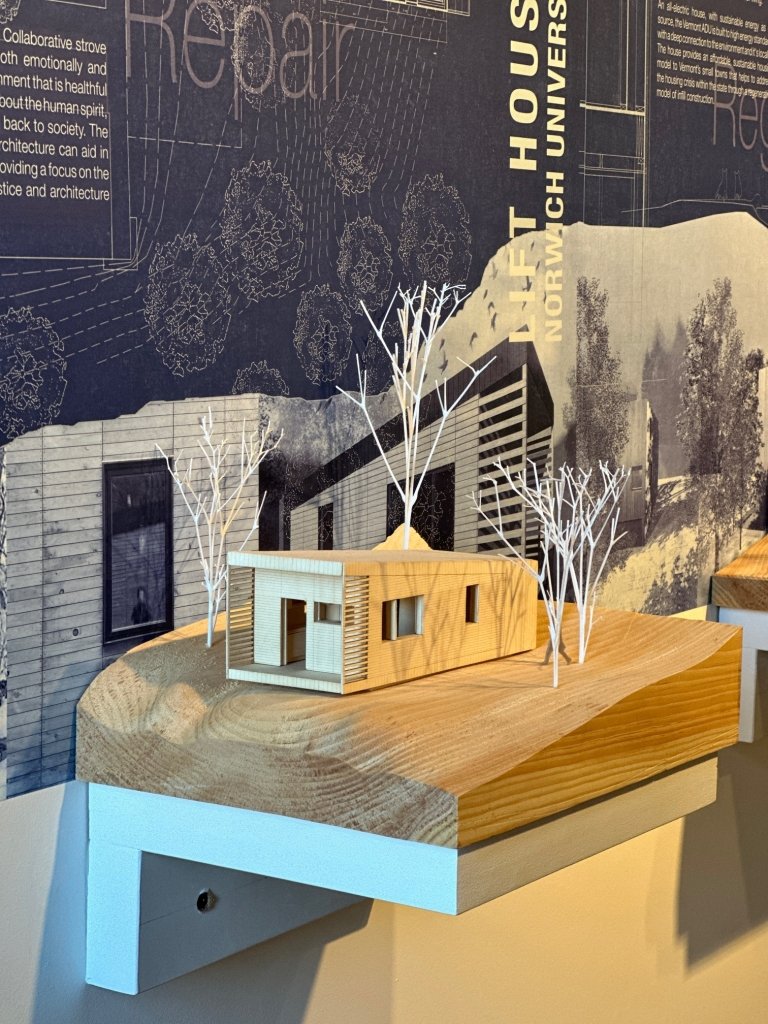 Architectural model featuring a detailed scale model of a minimalist house with surrounding trees displayed on a wooden platform, against a background of informational panels with text and diagrams.