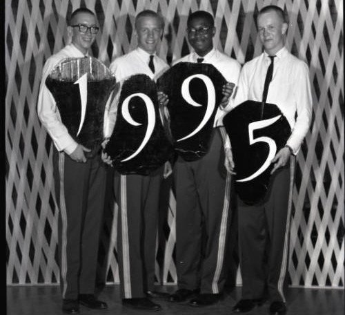 Black-and-white photograph of a group of Norwich University students with decorations from Junior Week for the Class of 1965 ; one of the decorations has been repositioned to read "1995" instead of 1965. In 1964, Junior Week was held from May 8 to May 10.